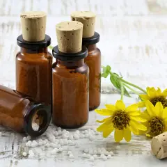 Bursztynowe buteleczki z korkami i białymi kuleczkami, symbolizujące homeopatię, obok żółtych kwiatów na drewnianym tle.