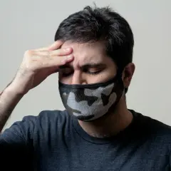 Mężczyzna z maską na twarzy trzyma rękę na czole, cierpiąc z powodu bólu głowy, który może być objawem autoimmunologicznego zapalenia mózgu.