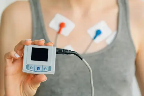 Dłoń trzyma urządzenie do monitorowania ekg serca. Na klatce piersiowej widoczne elektrody.