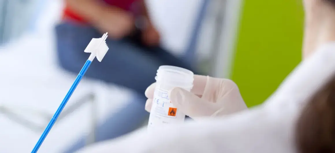 Ręka w rękawiczce trzyma pojemnik na próbkę do cytologii płynnej. W tle widać pacjentkę.