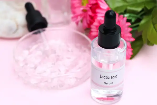 Serum z kwasem mlekowym w szklanej buteleczce z pipetą, obok różowych kwiatów.